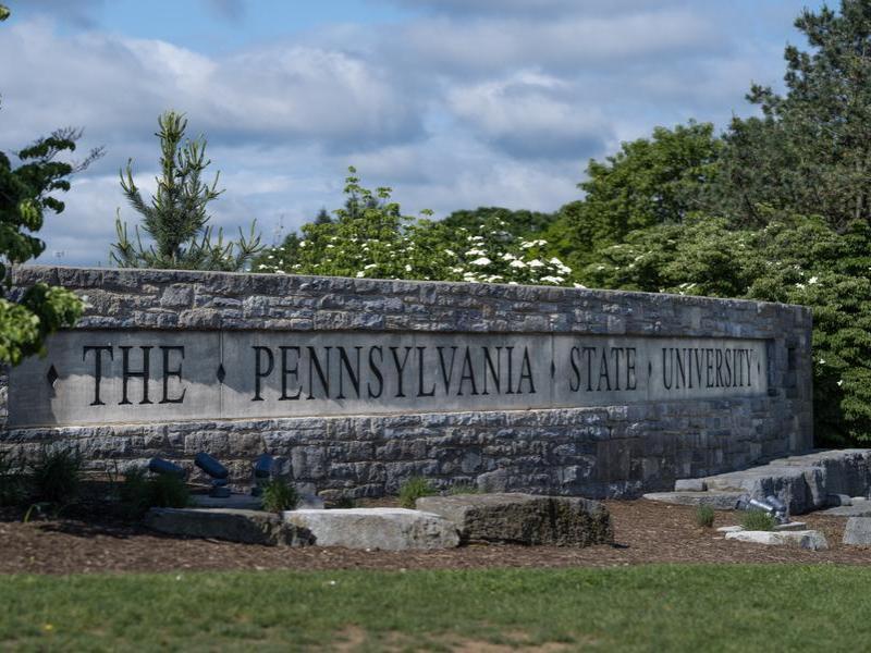 Image of "The Pennsylvania State University" sign at Penn State's University Park campus with a background of greenery and clouds.