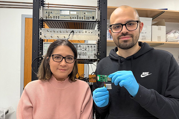 Two people wearing glasses pose in a lab. One holds an electronic sensing device.