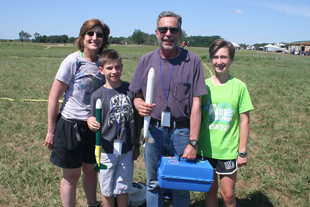 A family portrait of two adults and two kids standing in a field holding model rockets. 