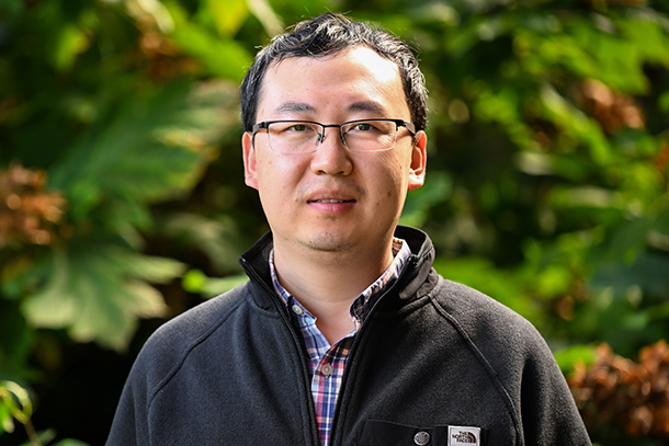 An individual poses outdoors in front of trees.