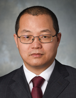 A person wearing glasses and business professional clothing poses for a photo in front of a grey background.